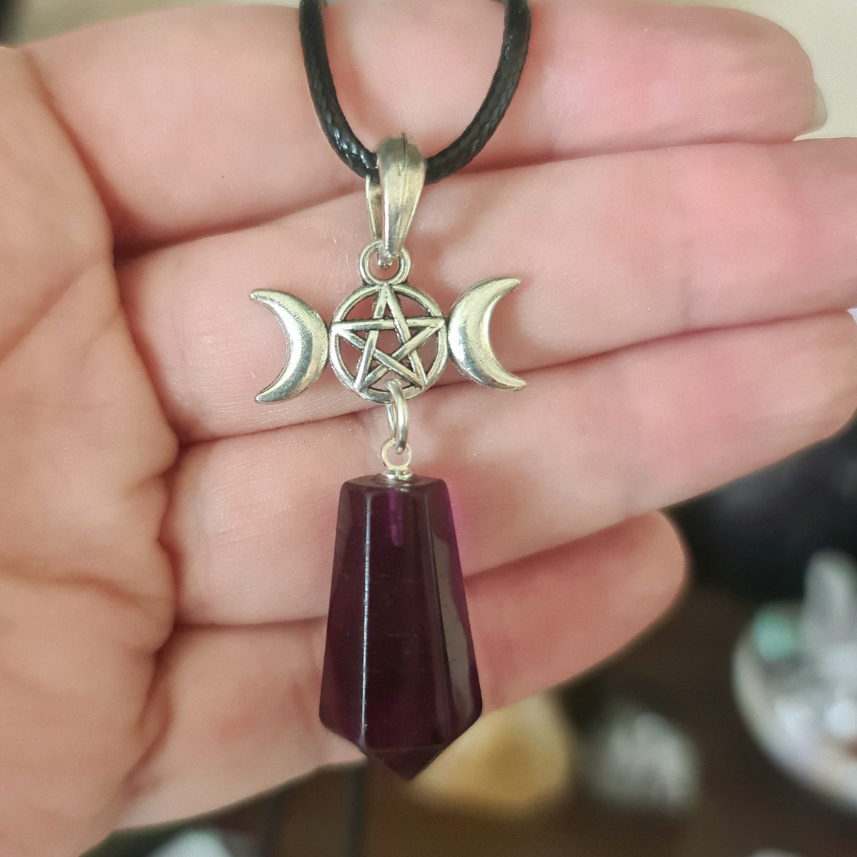 A Fluorite Triple Moon Goddess Necklace with a purple Fluorite pendant and silver-colored metal charm, held in a person's hand.