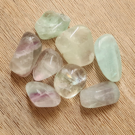 A collection of polished Fluorite tumble stones in various shades of purple, green, and transparent, arranged on a wooden surface.