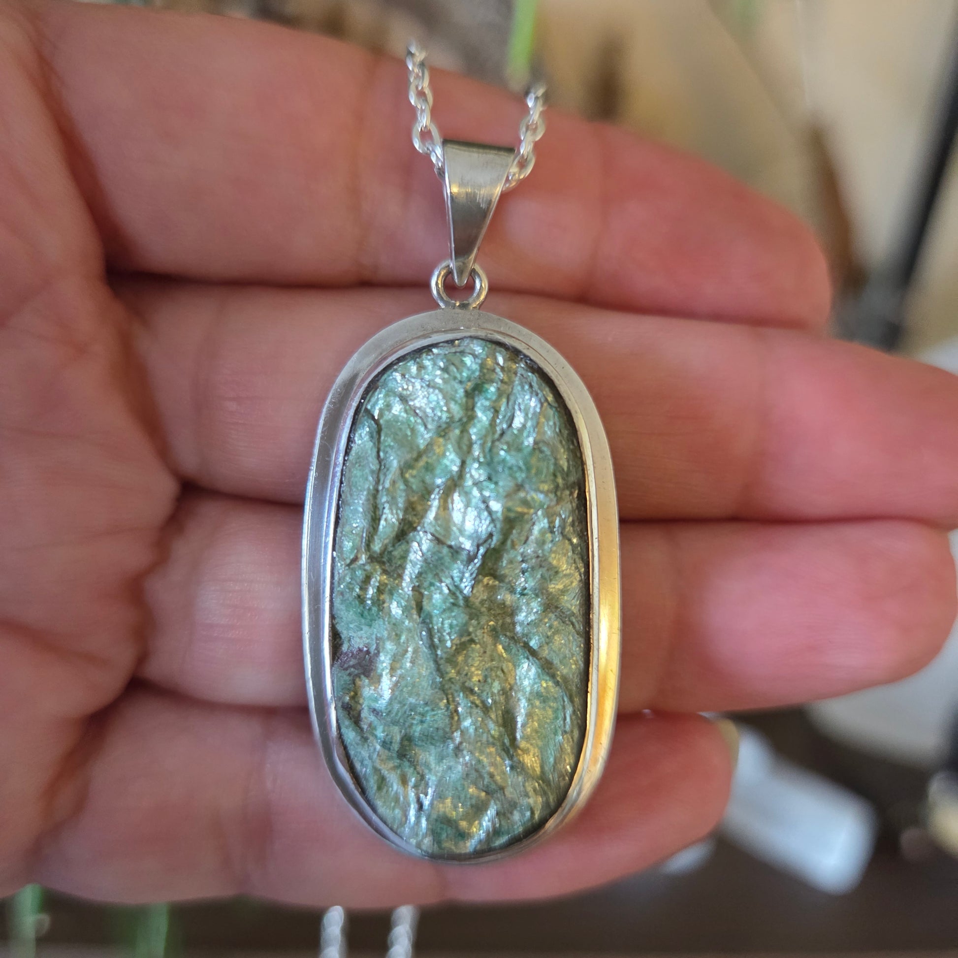 A hand holding a silver pendant with a green mica stone on a necklace.