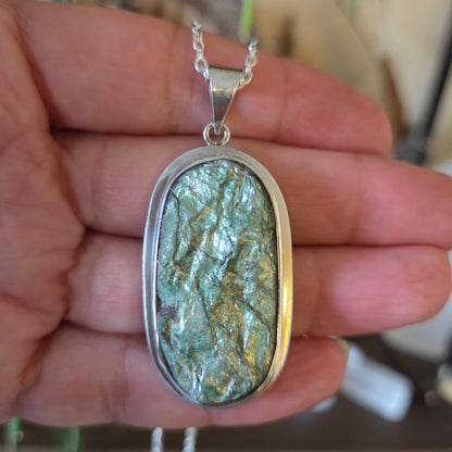 A hand holding a silver pendant with a green mica stone on a necklace.