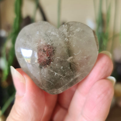 A hand holding a gray garden quartz crystal with a heart-like shape, with natural mineral inclusions.