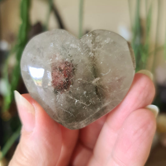 A hand holding a gray garden quartz crystal with a heart-like shape, with natural mineral inclusions.