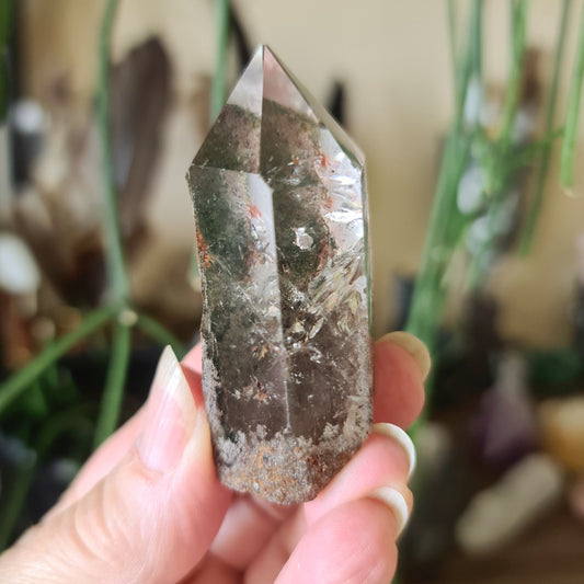 A hand holding a polished piece of garden quartz with a rough, natural edge, against a backdrop of similar crystals and plant elements.