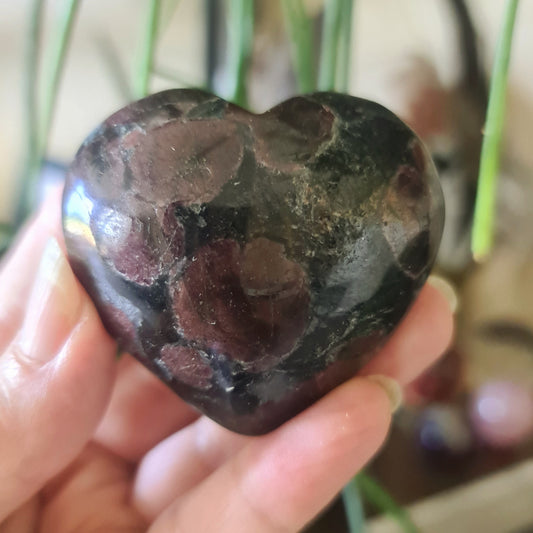 A hand holding a polished garnet crystal with a heart-like shape, with a blurred green background.