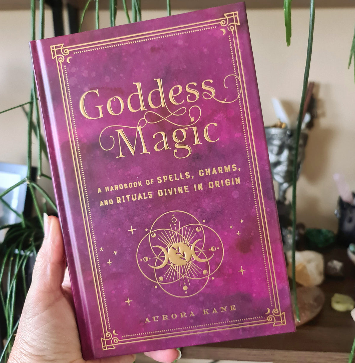 A hand holding a purple hardback book titled 'GODDESS MAGIC' with a golden circular emblem on the cover.