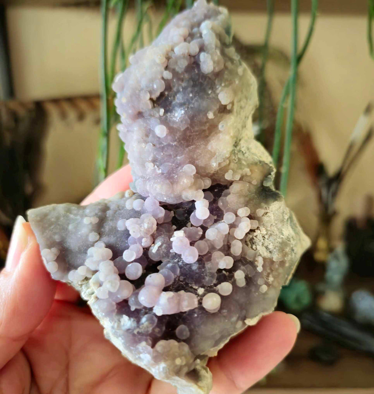 A close-up of a Grape Agate cluster held in a person's hand, with a blurred background containing other decorative stones.