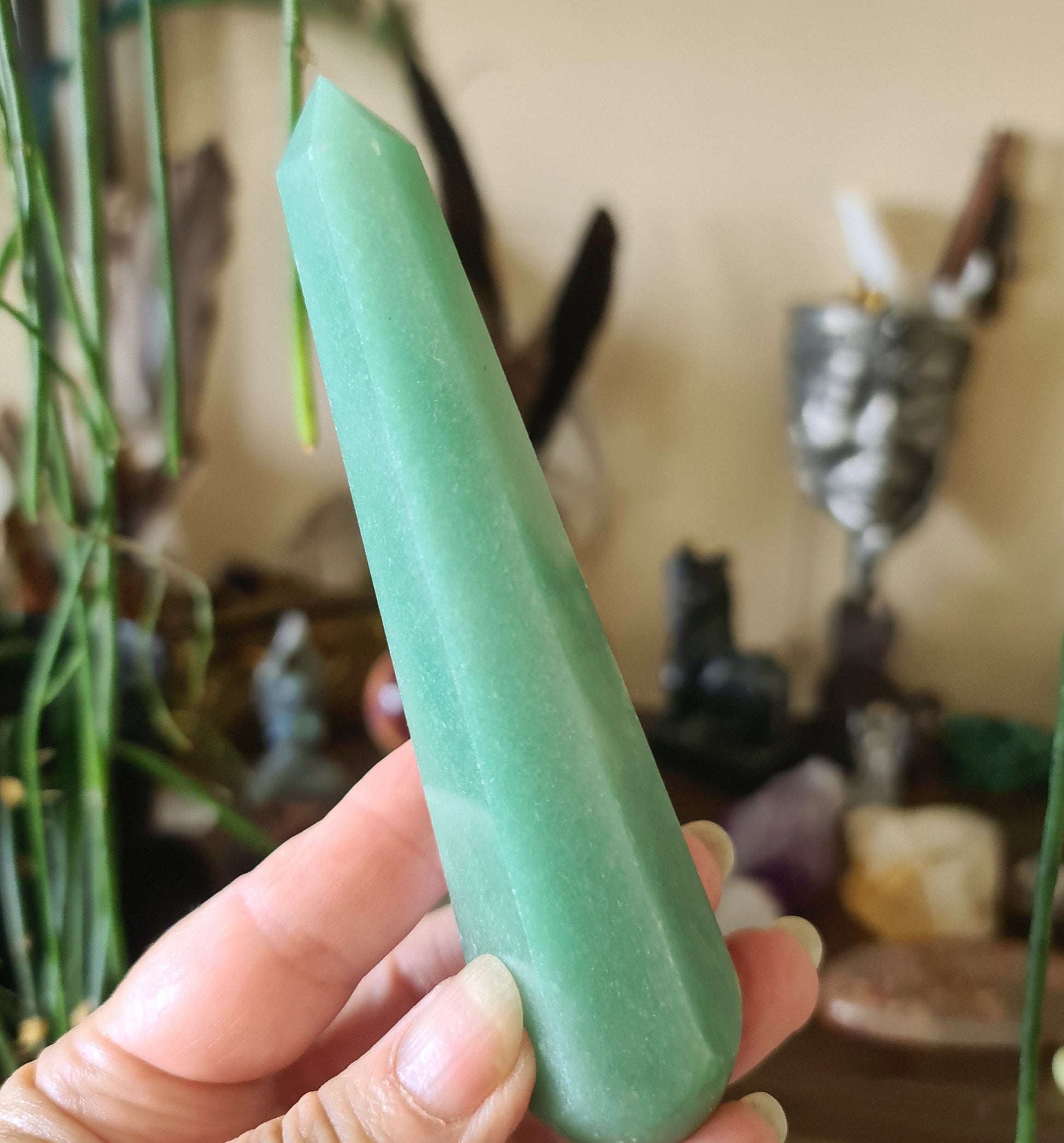 A green aventurine crystal wand held in a person's hand, with a blurred background featuring various crystal figures and objects.