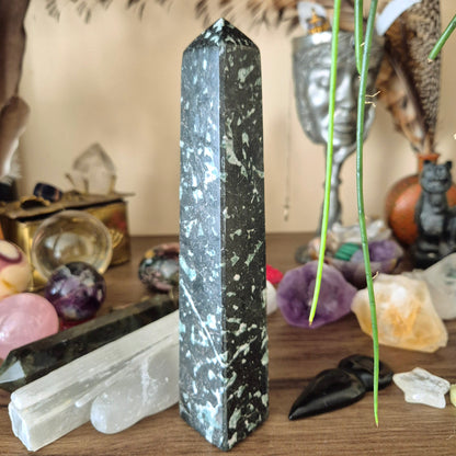 A green jasper obelisk standing upright on a wooden surface, surrounded by various crystal and decorative items.