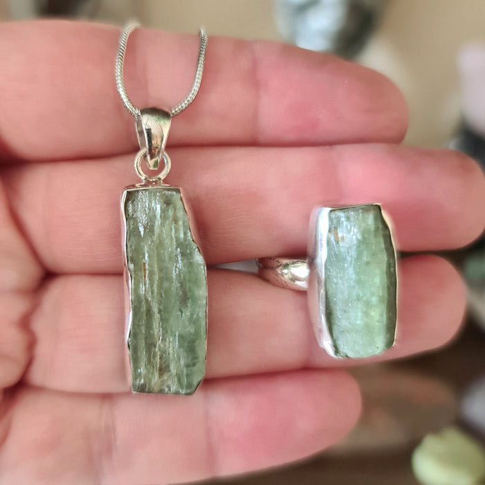 A green kyanite crystal pendant and ring set, with the pendant being held by a hand and the ring on a finger.