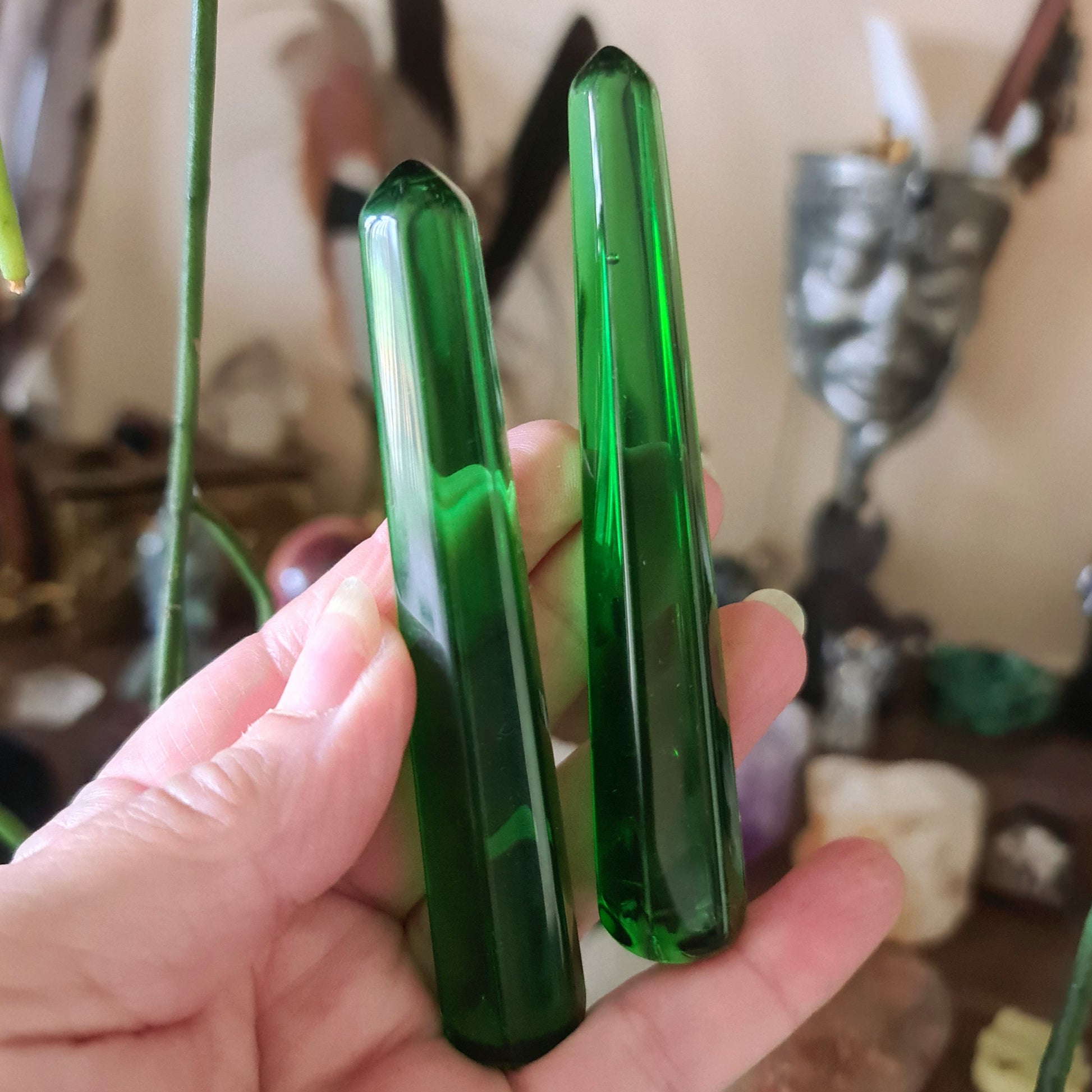 Two green obsidian wands being held, with a blurred background featuring various crystal items.
