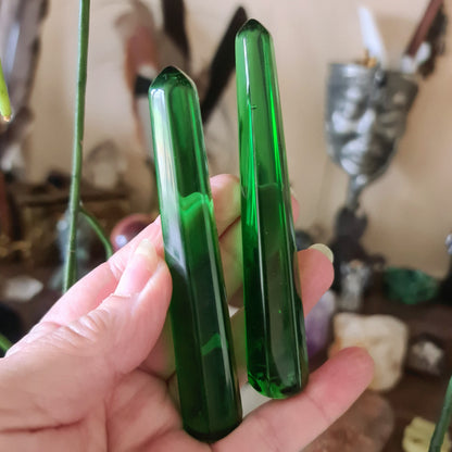 Two green obsidian wands being held, with a blurred background featuring various crystal items.