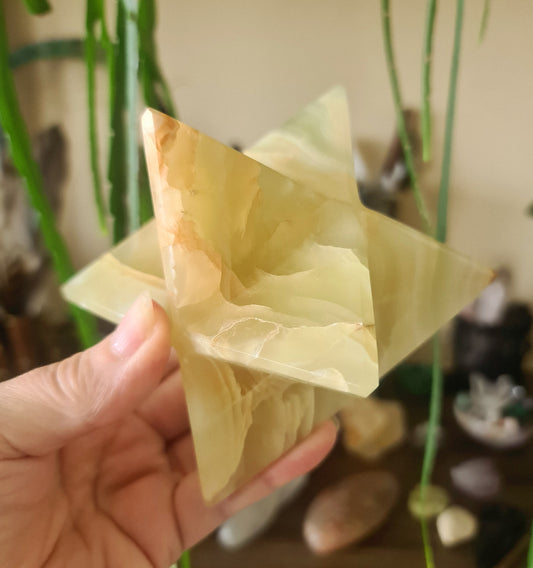 A green onyx merkaba, held in a person's hand, with a triangular pyramidal shape and a polished, smooth surface.