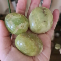 Three polished green opal palm stones held in a person's hand, with a blurred background suggesting a soft-focus effect.