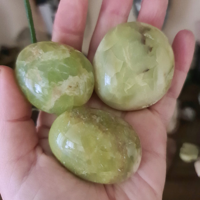 Three polished green opal palm stones held in a person's hand, with a blurred background suggesting a soft-focus effect.