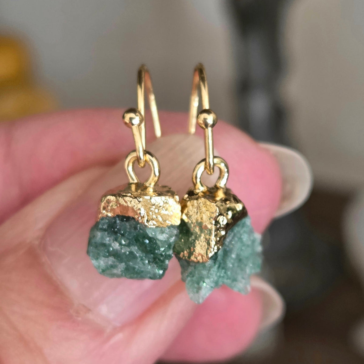 A close-up of a person's hand holding a pair of gold drop earrings with rough green crystal pendants.