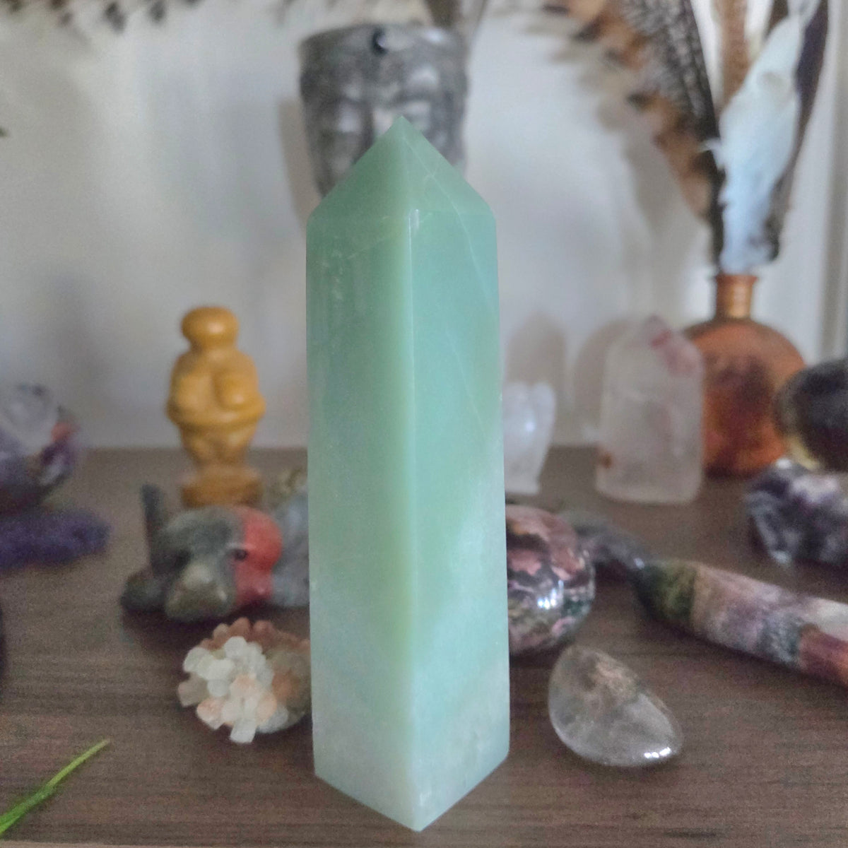 A light green Aventurine crystal tower stands on a wooden surface surrounded by various polished stones and crystals in a softly lit setting.
