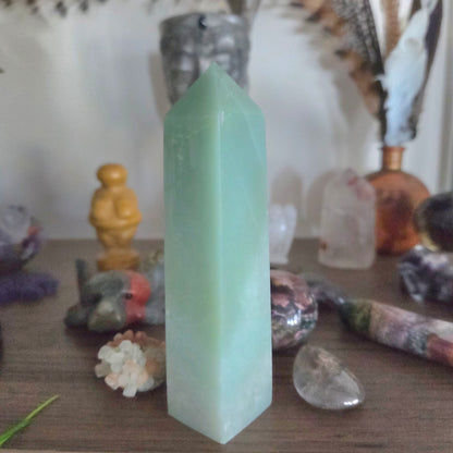 A light green Aventurine crystal tower stands on a wooden surface surrounded by various polished stones and crystals in a softly lit setting.