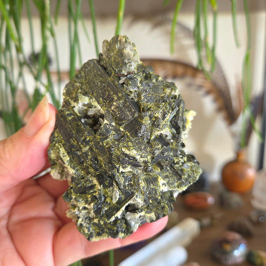 Hand holding a large green mineral crystal with a blurred indoor setting