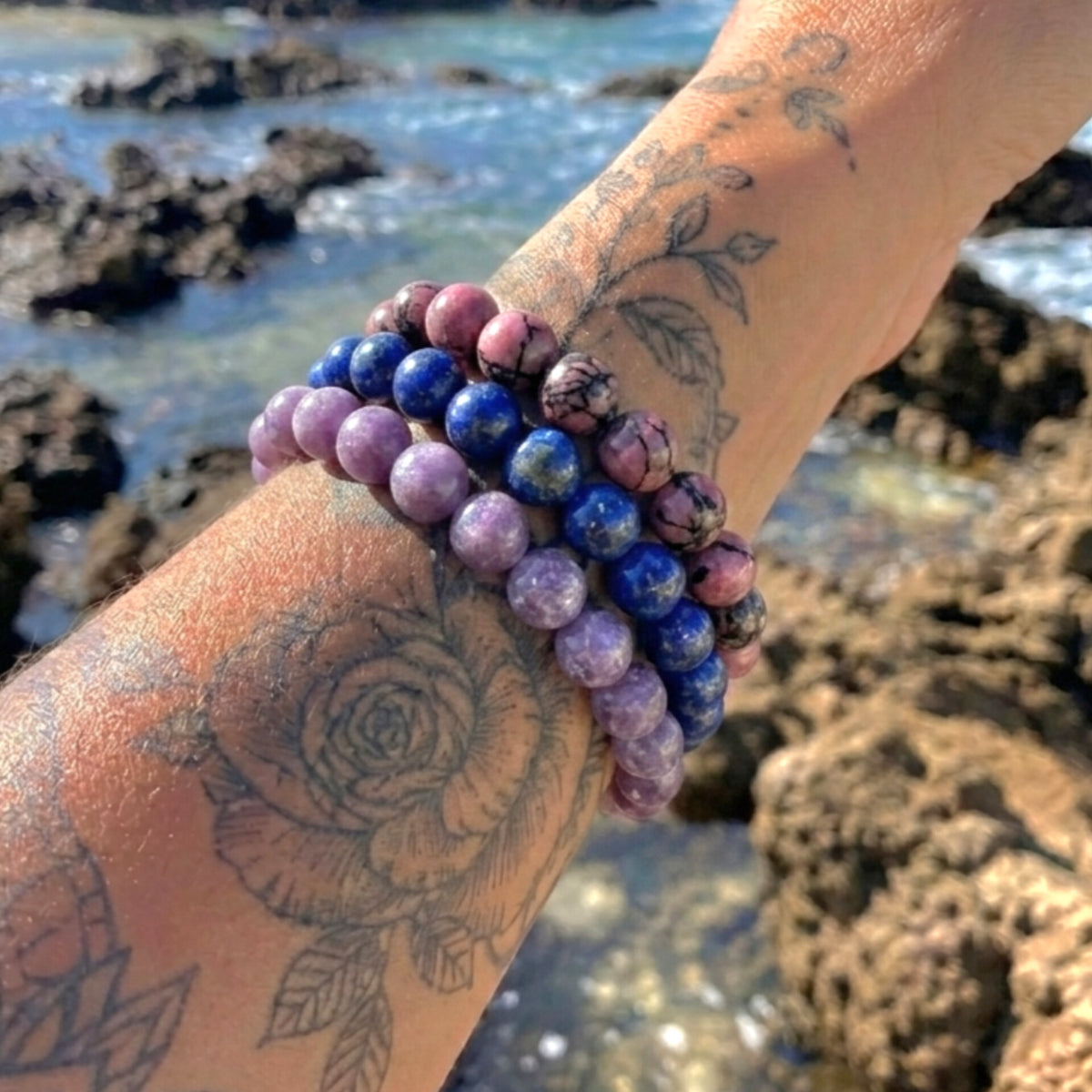 A close-up of a tattooed wrist wearing three beaded bracelets outdoors by the rocky ocean shore. The bracelets include round purple and blue marbled beads, with one bracelet featuring pink and purple marbled beads.