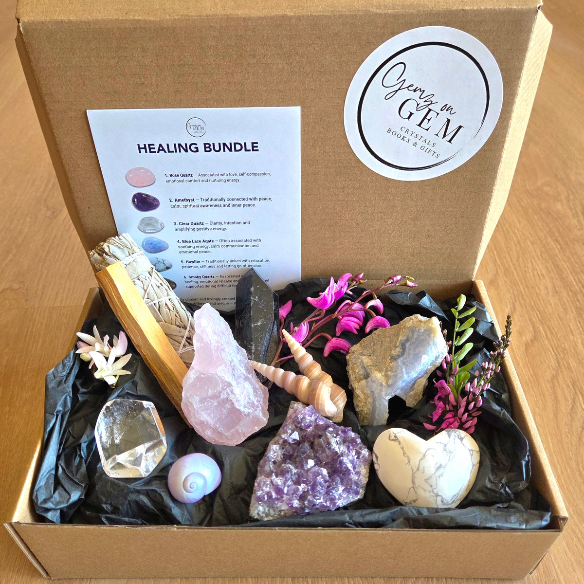 A wooden gift box opened to reveal a curated crystal and gemstone bundle on black tissue paper. The set includes a pink quartz crystal, a purple amethyst cluster, a white marble heart stone, a clear faceted crystal, a spiral shell, a black pointed crystal, dried white sage, a wooden wand, and small pink and purple flowers.