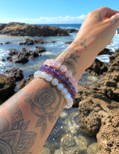 A close-up of a tattooed forearm and hand wearing three beaded bracelets outdoors by the rocky ocean shore. The bracelets include light pink, deep purple, and milky white iridescent beads.