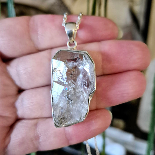 A hand holding a silver Herkimer diamond pendant with a chain, showcasing the crystal's clarity and inclusions.
