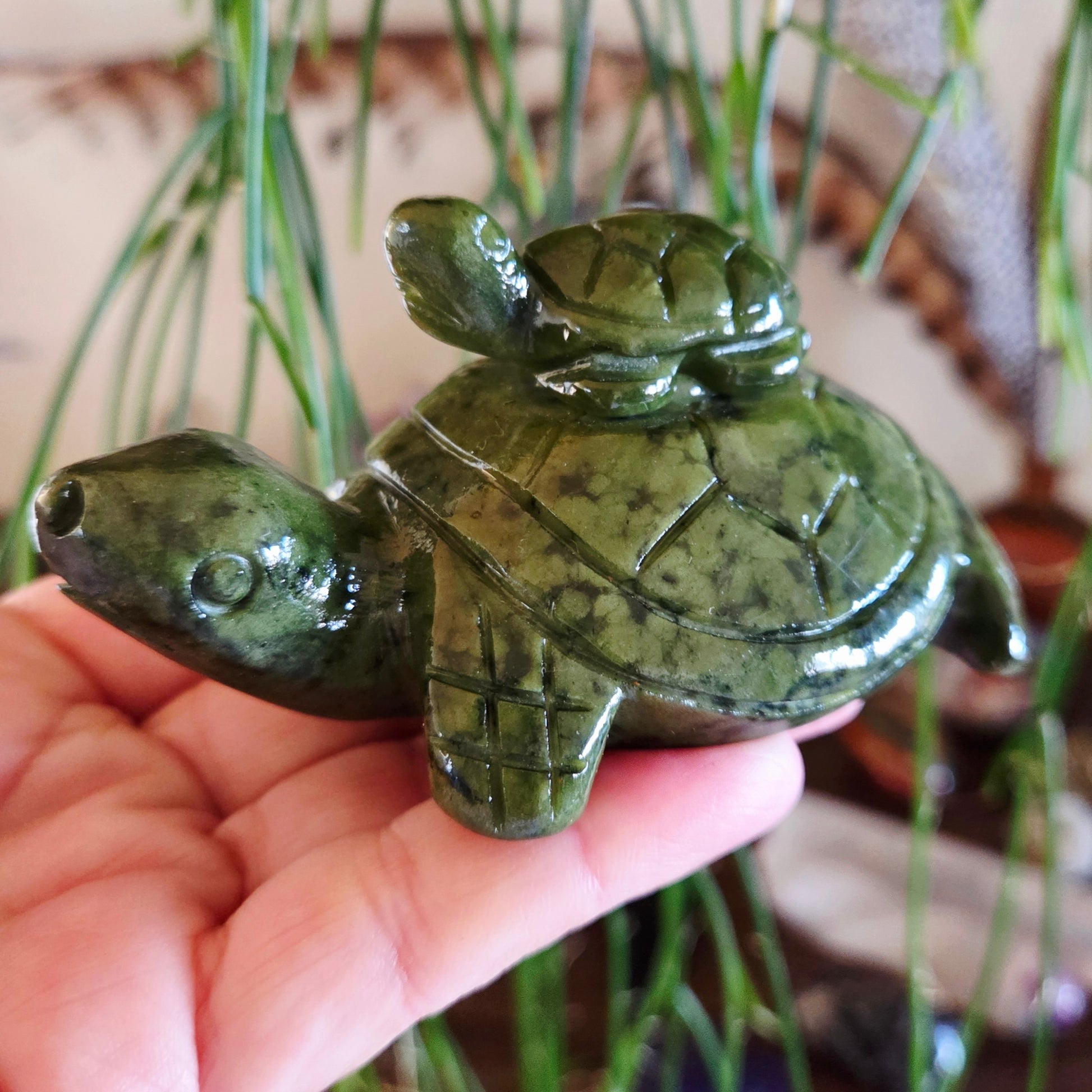 A hand holding a green jade figurine of a mother turtle with a baby turtle on its back, placed against a backdrop with green plant leaves.