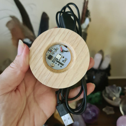 A person holding a round LED light base with a timber (wooden) base and an attached USB cable.