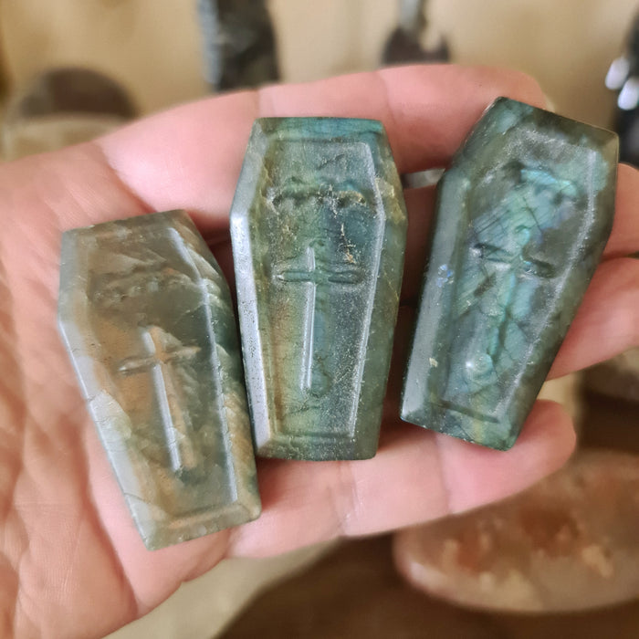 Three pieces of green Labradorite stone shaped like coffins, held in a person's hand against a backdrop of other stones.