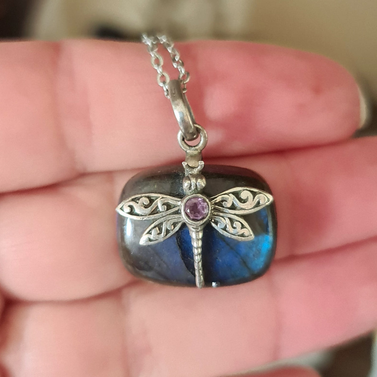 A labradorite dragonfly necklace with an amethyst crystal set in 925 sterling silver, held in a person's hand.