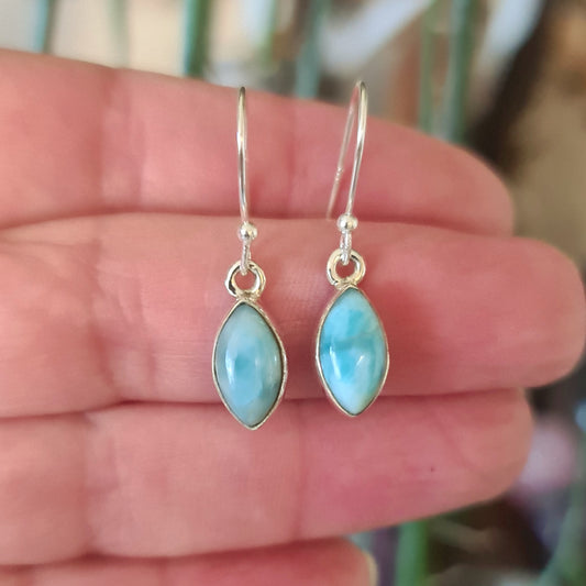 A pair of silver earrings with a large blue larimar stone and smaller clear crystal accents, held in a hand against a blurred green background.