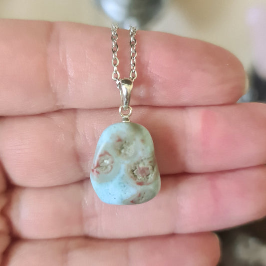 A blue Larimar tumble pendant on a silver plated chain, held in a person's hand.