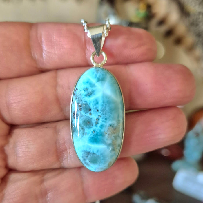 An oval-shaped Larimar pendant set in a silver mounting, held by a hand.
