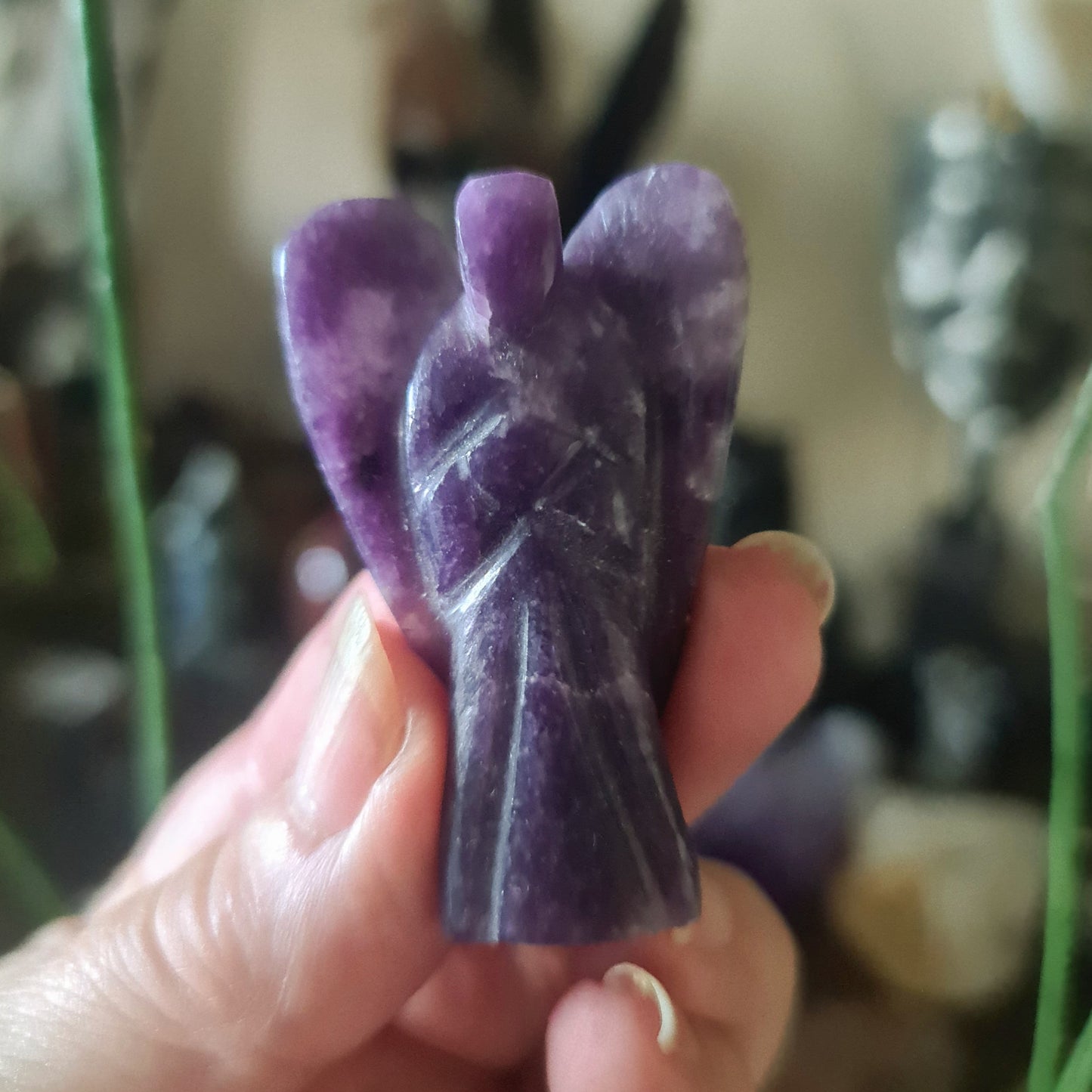 A hand holding a carved purple Lepidolite crystal in the shape of an angel.