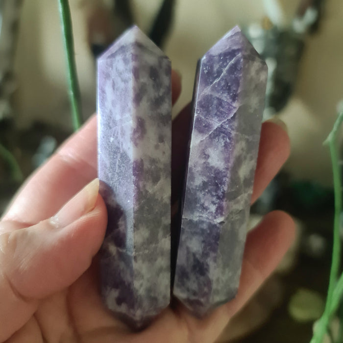 A hand holding a double-pointed Lepidolite crystal with a purplish hue.