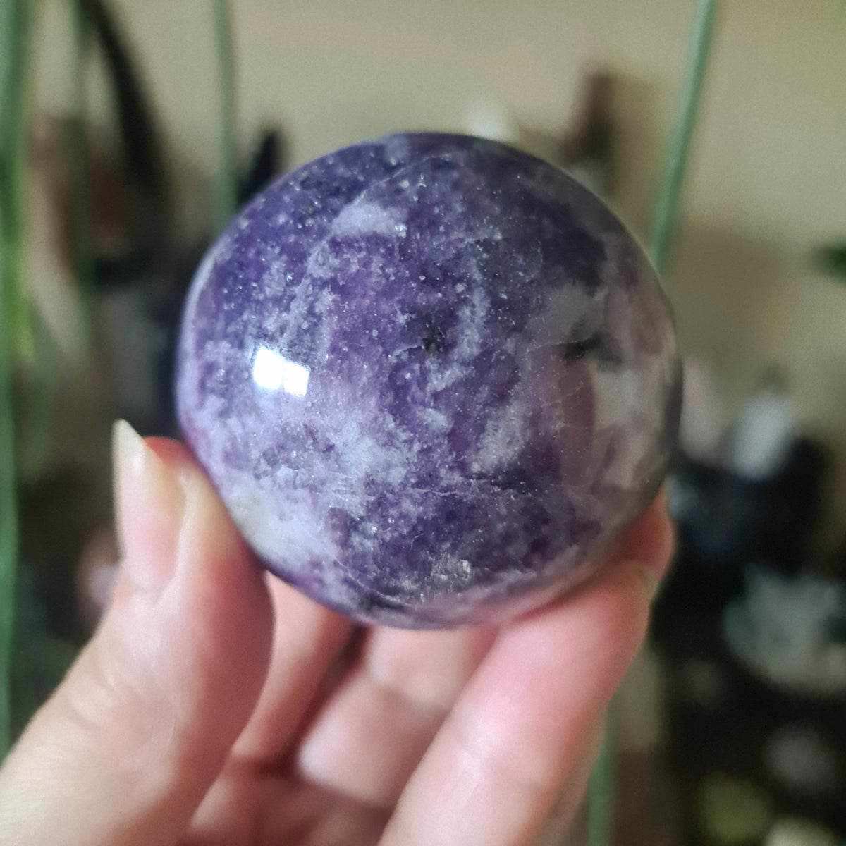 A purple Lepidolite crystal sphere held in a person's hand, with a blurred background suggesting indoor setting