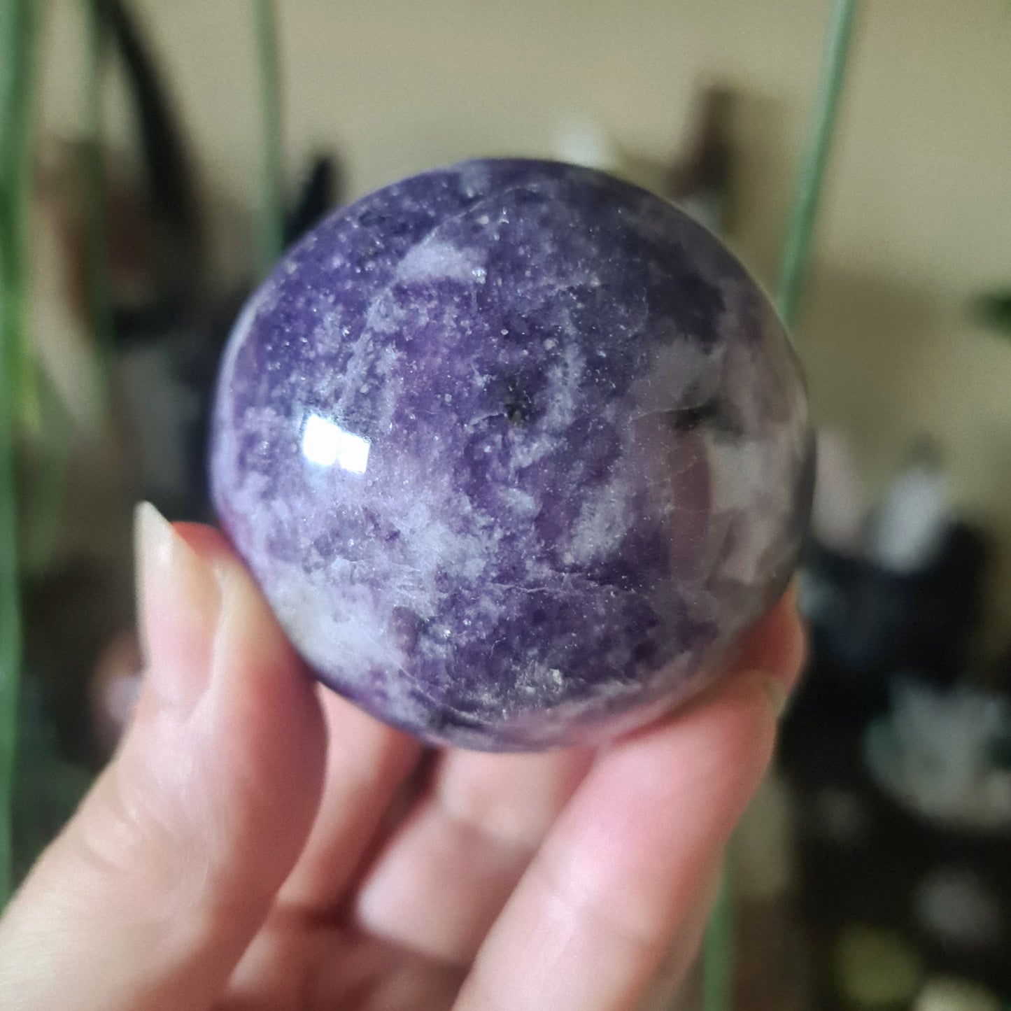 A purple Lepidolite crystal sphere held in a person's hand, with a blurred background suggesting indoor setting