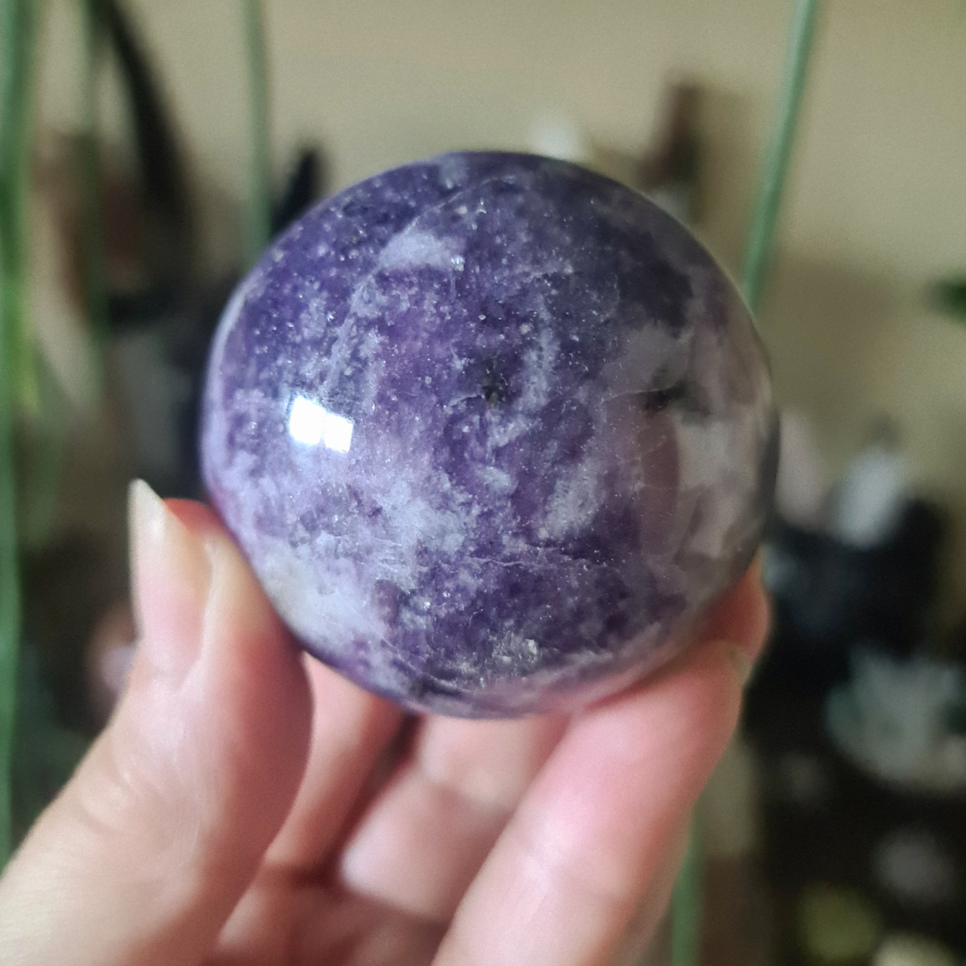 A purple Lepidolite crystal sphere held in a person's hand, with a blurred background suggesting indoor setting