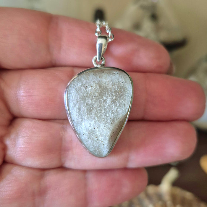 A silver-colored pendant featuring a piece of desert glass, held in a person's hand.