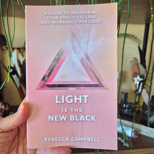 This image features a book titled "Light Is The New Black" by Rebecca Campbell. The cover, set against a soft pink background, showcases a minimalist design with a prominent, geometric triangle in white and pink, symbolizing perhaps the author's spiritual theme or the book's title.