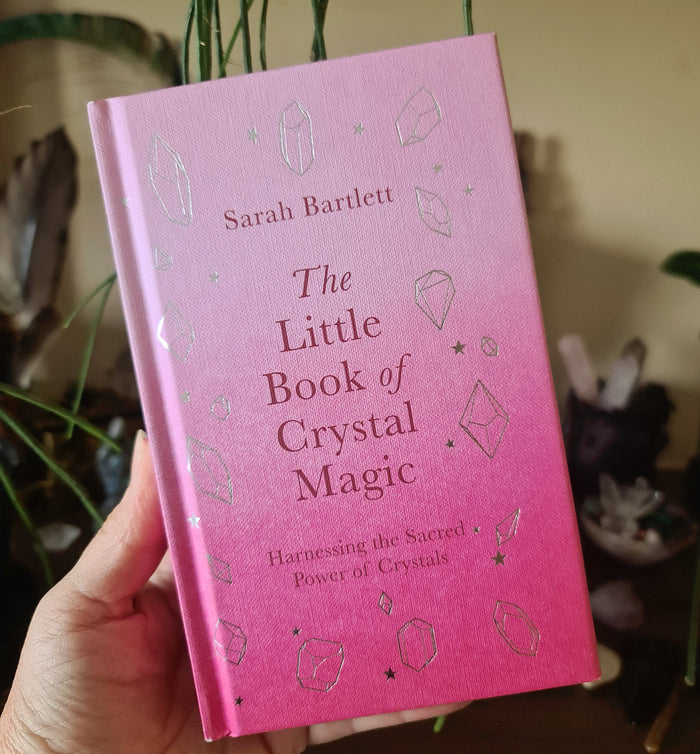 A hand holding a pink book titled 'The Little Book of Crystal Magic' with crystal-themed illustrations on the cover.
