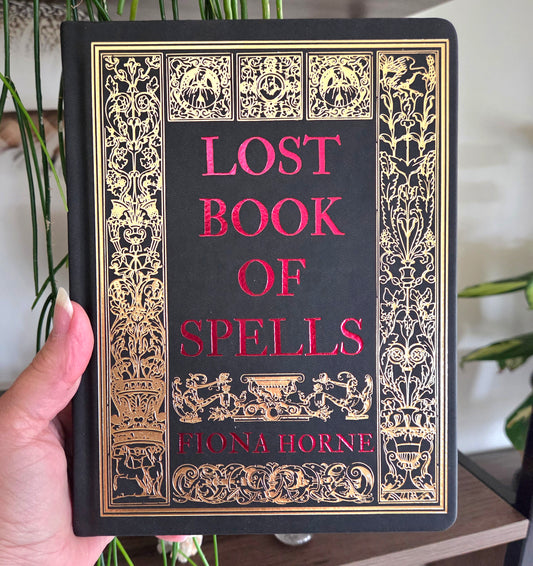 A hand holds a hardcover book titled 'Lost Book of Spells' by Fiona Horne. The book cover is black with ornate gold filigree borders and the title and author name in red text.