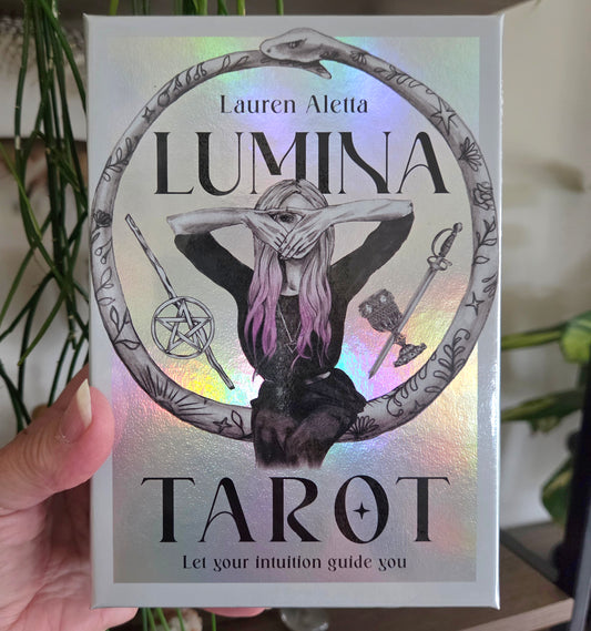 A hand holds a silver holographic box of the 'Lumina Tarot' deck by Lauren Aletta. The box features a black and white illustration of a woman with pink hair inside a circular frame, surrounded by occult symbols, with a rainbow iridescent sheen.