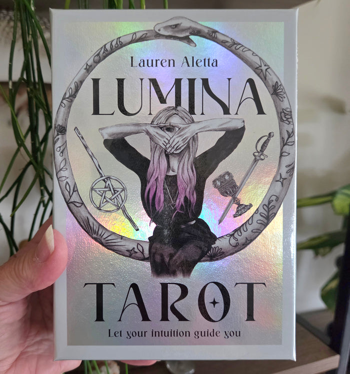 A hand holds a silver holographic box of the 'Lumina Tarot' deck by Lauren Aletta. The box features a black and white illustration of a woman with pink hair inside a circular frame, surrounded by occult symbols, with a rainbow iridescent sheen.