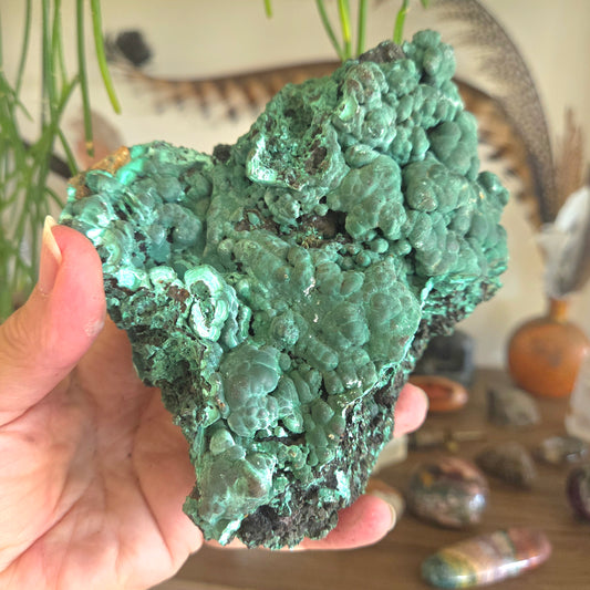 Hand holding a large green mineral specimen with a blurred indoor setting