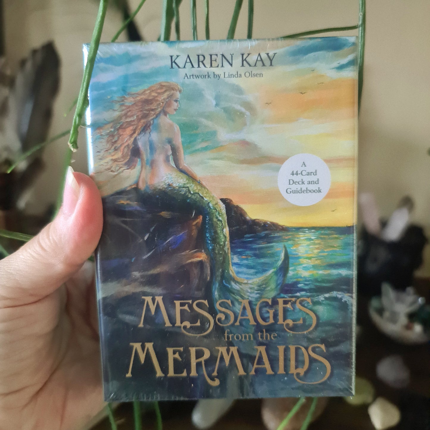 A hand holding a card from the 'Messages From The Mermaids' Oracle Deck, featuring artwork of a mermaid sitting on a rock under a sunset sky.