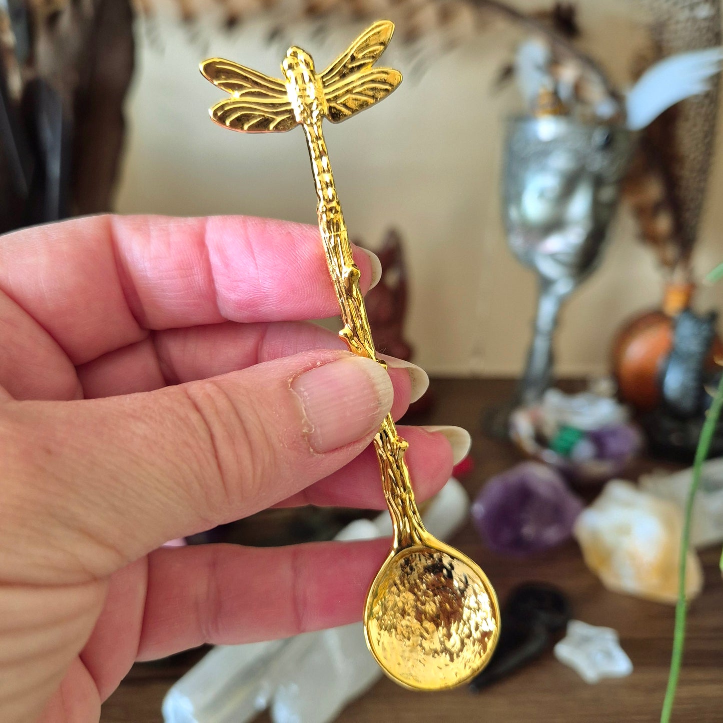 A gold-colored metal spoon with a dragonfly design on the handle, held in a person's hand.
