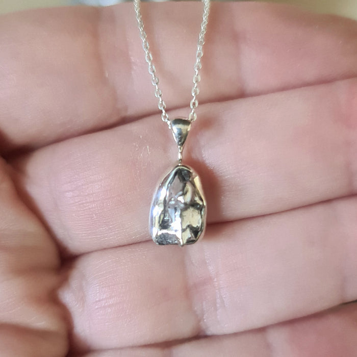 A polished meteorite pendant set in a sterling silver setting on a sterling silver chain, held in a person's hand.