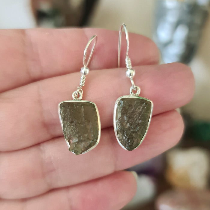 A pair of silver earrings featuring moldavite crystals, held in a person's hand.