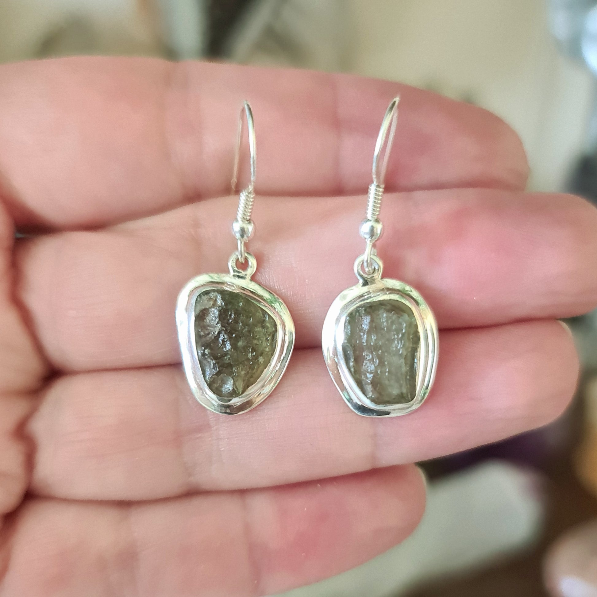 A pair of silver earrings with moldavite stones set in them, held in a hand.
